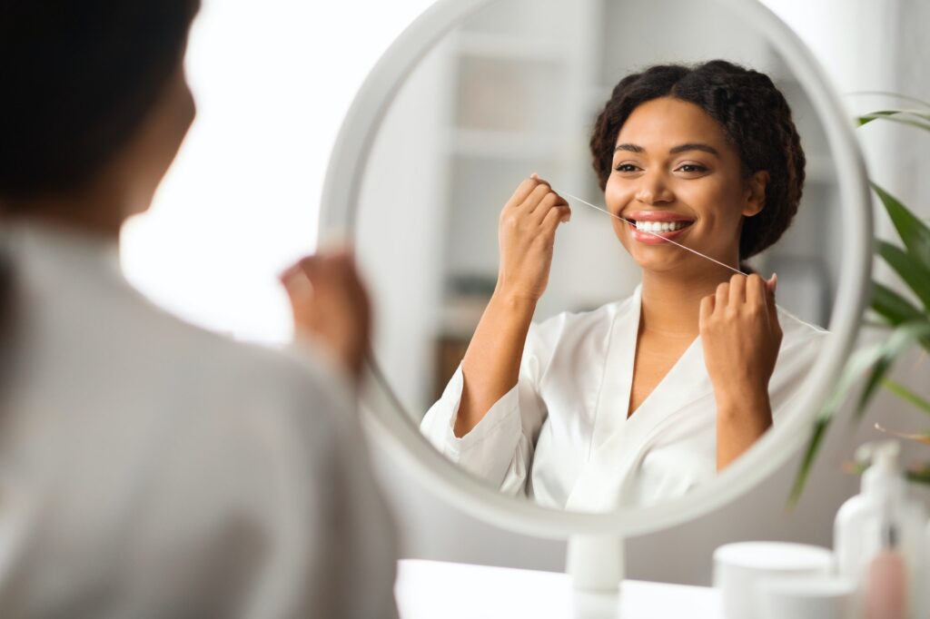 Smiling Black Woman Using Dental Floss Near Mirror At Home